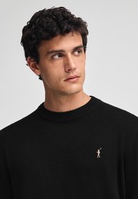 Maglione nero a maglia con scollo rotondo, caratterizzato da un piccolo logo rosa sul lato sinistro del petto. Il tessuto ha una consistenza morbida e liscia.