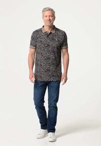 Donker bloemenpatroon polo shirt met gestreepte kraag, gecombineerd met blauwe jeans en witte sneakers. Model staat met de handen langs het lichaam.