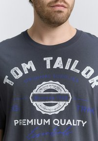 Czarny bawełniany t-shirt w kolorze ciemnoszarym z białym i niebieskim nadrukiem, logo w stylu vintage oraz okrągłym dekoltem. Krótkie rękawy z miękkiej tekstury.