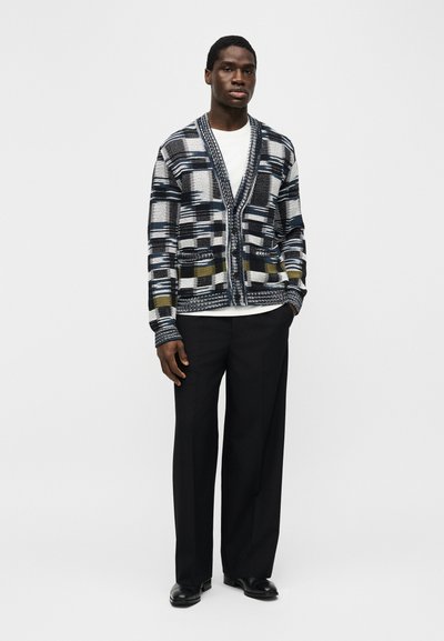Νέος άνδρας που στέκεται φορώντας ένα πλεκτό ζακέτο με σχέδια σε μαύρο, άσπρο και ελιά, λευκό πουκάμισο, μαύρο παντελόνι με φαρδιά πόδια και μαύρα παπούτσια.