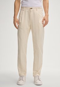 Beige, op maat gemaakte broek met een platte voorkant, zijzakken en een enkele knoopsluiting. Draagt wit sneakerpaar. Glad katoenen stof.