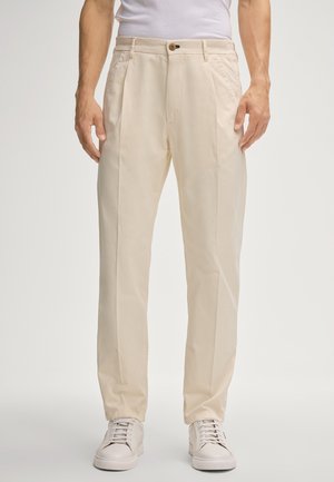 Beige, op maat gemaakte broek met een platte voorkant, zijzakken en een enkele knoopsluiting. Draagt wit sneakerpaar. Glad katoenen stof.