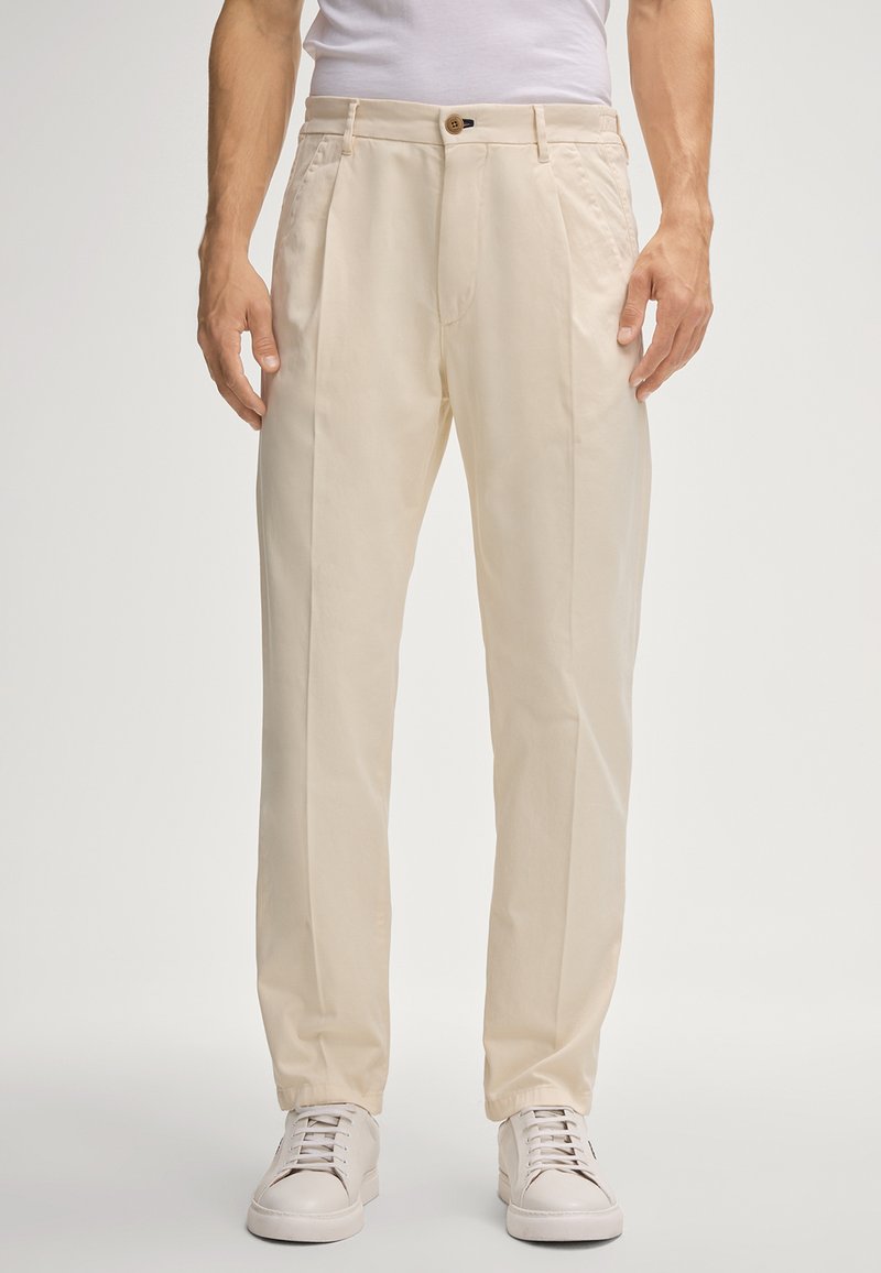 Beige, op maat gemaakte broek met een platte voorkant, zijzakken en een enkele knoopsluiting. Draagt wit sneakerpaar. Glad katoenen stof.