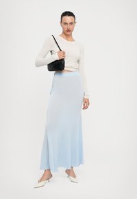 Saia longa azul clara com uma textura suave, apresentando babados laterais. Combinada com uma blusa justa de manga longa branca e uma mala preta.