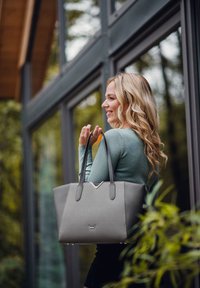 Graue Tasche aus glattem synthetischem Material, mit einem V-förmigen Designelement, zwei schwarzen Griffen und einem dezenten Logo auf der Vorderseite.