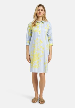 Frau trägt ein knielanges blaues Kleid mit weißen vertikalen Streifen und gelben Blumenmustern, kombiniert mit hellbraunen Sandalen, steht frontal.