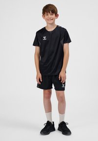 Pojke som står och ler, iklädd svart sport-T-shirt och shorts, vita strumpor och svarta sneakers mot en helt vit bakgrund.