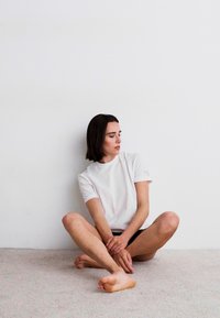 White short-sleeve t-shirt with a round neckline, paired with black shorts. Model sits on a beige carpet against a white wall.