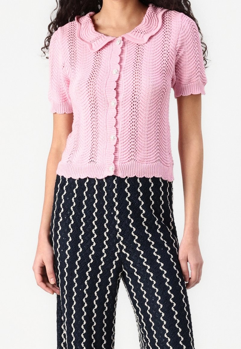 Růžový pletený cardigan se zvlněným límcem, krátkými rukávy a zapínáním na knoflík, v kombinaci s kalhotami s námořnickým zigzag vzorem.