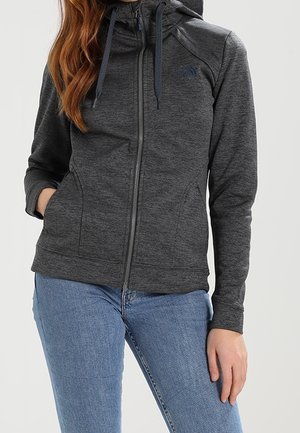 Femme portant un sweat à capuche zippé gris foncé avec une capuche et un jean bleu, debout, une main dans la poche et l'autre détendue de chaque côté.