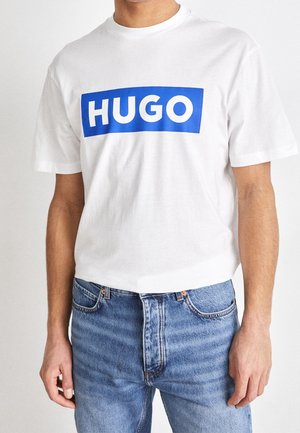 Mężczyzna ubrany w białą koszulkę z krótkim rękawem z niebieskim prostokątnym logo z napisem "HUGO" oraz jasnoniebieskie jeansy.