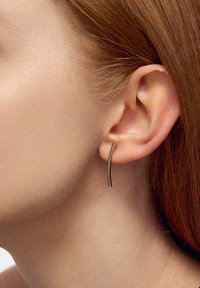 Złote, zakrzywione kolczyki o eleganckim, minimalistycznym designie, wieszane tuż poniżej płatka ucha. Gładka faktura, smukły profil i wypolerowane wykończenie.