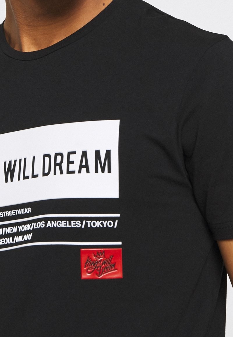 Camiseta de algodón negra con un diseño de bloques blancos que incluye el texto "WILL DREAM" y una etiqueta de marca roja con un logo de corona en el dobladillo.
