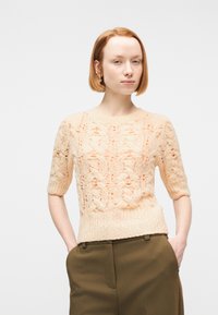 Νεαρή γυναίκα με κοντά κόκκινα μαλλιά, φορώντας ροδακινί κασμιρένιο πουλόβερ με σύντομο μανίκι και ψηλόμεσο παντελόνι σε ελαιί απόχρωση, με τα χέρια στις τσέπες.