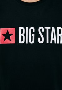 Fekete ing fehér "BIG STAR" felirattal, a mellkason egy piros négyzetben fekete csillag szimbólummal.