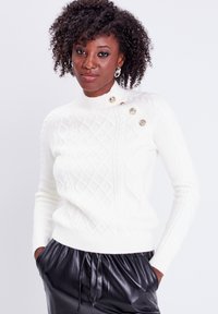 Suéter blanco de punto con un cuello alto, patrón de diamante y acentos de botones dorados en el hombro, combinado con pantalones de cuero negros.