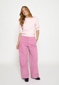 Lyserød strikket trøje med korte pufærmer, kombineret med lyserøde bukser med vide ben. Bukserne har talje i midjeniveau og to forlommer.