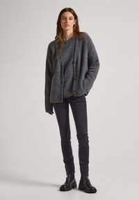Cárdigan gris de punto con detalles de botones, mangas texturizadas y dobladillo acanalado, combinado con jeans oscuros y botas negras hasta el tobillo.