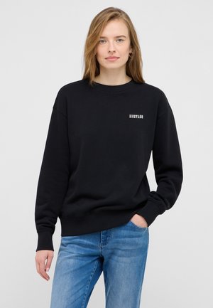 Black sweatshirt with ribbed texture, round neck, and dropped shoulders. Features "Mustang" logo in light gray on the front. Worn with blue jeans.