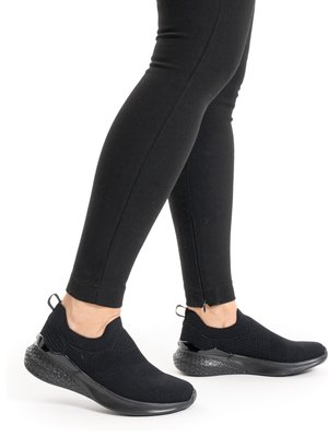 Person wearing black slip-on athletic shoes and black fitted pants, standing with one foot slightly forward on white background.