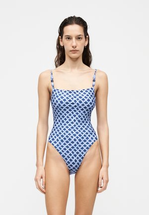Femme debout face à l’avant portant un maillot de bain une pièce à motifs bleus et blancs avec fines bretelles et détails perlés.