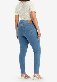 Jeans de mezclilla azul de talle alto con un ajuste slim, mostrando los bolsillos traseros y la etiqueta de la marca. Se combinan con una blusa de manga corta blanca texturizada y sandalias.