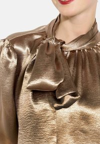 Kobieta w błyszczącej, miedzianej satynowej bluzce z dużą kokardą zawiązaną na szyi, uśmiechająca się z jaskrawoczerwoną szminką.