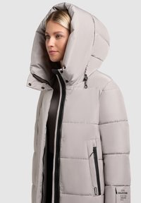 Giacca puffer grigia chiara con un grande cappuccio, chiusura frontale con zip e tasche laterali. Presenta un motivo trapuntato e dettagli con zip nera.
