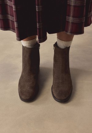 Braune Wildleder-Stiefeletten mit seitlichen Reißverschlüssen, getragen mit weißen Socken und einem karierten Rock auf einem beigen Boden.