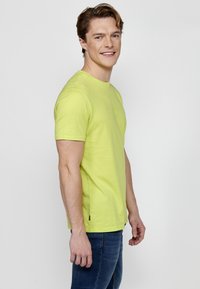 Cămașă scurtă de culoare verde lime, confecționată dintr-un material moale, cu guler rotund, croială dreaptă și ventilație laterală pentru libertatea de mișcare.