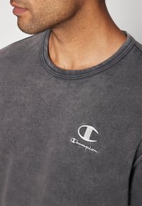 Bas du visage et cou d'un homme portant un sweatshirt Champion gris foncé délavé avec un logo blanc brodé sur le côté gauche de la poitrine.