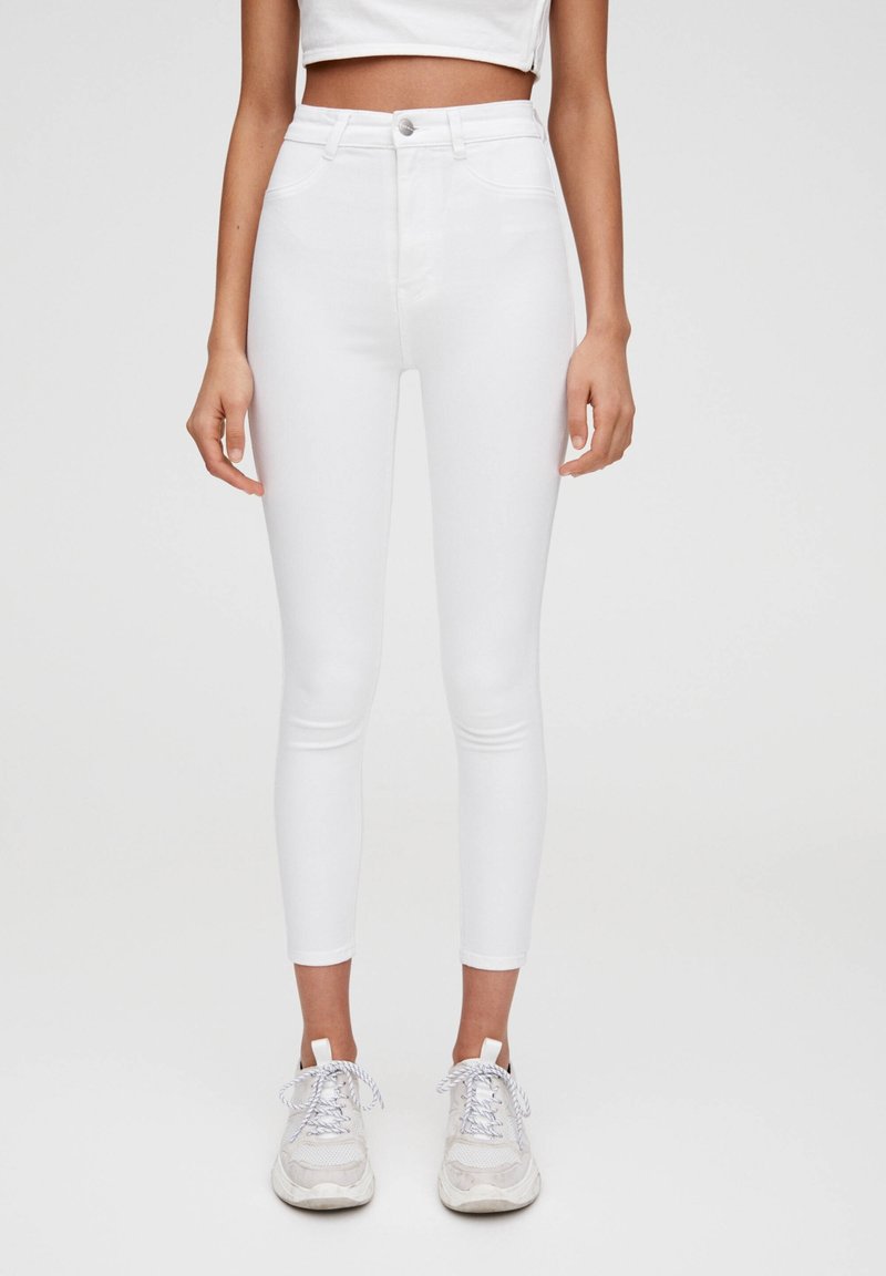 Femme portant un jean skinny blanc taille haute avec fermeture par bouton et zip, associé à des baskets blanches et un top court blanc.
