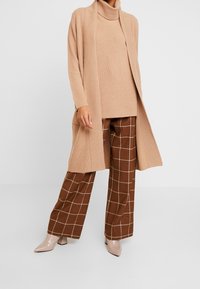 Beige ribget turtleneck sweater parret med brune ternede vidde bukser. Sættet kompletteres af nude hæle og en lang cardigan.