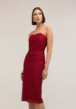 Frau in trägerlosem, knielangem roten Spitzenkleid mit Blumenmuster, trägt goldene Ohrringe und neutrales Make-up, steht vor schlichtem Hintergrund.