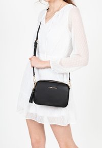 Borsa a tracolla in pelle nera con dettagli in oro, chiusura con zip e accentuazione a nappina. Texture liscia, forma rettangolare e logo del marchio visibile.