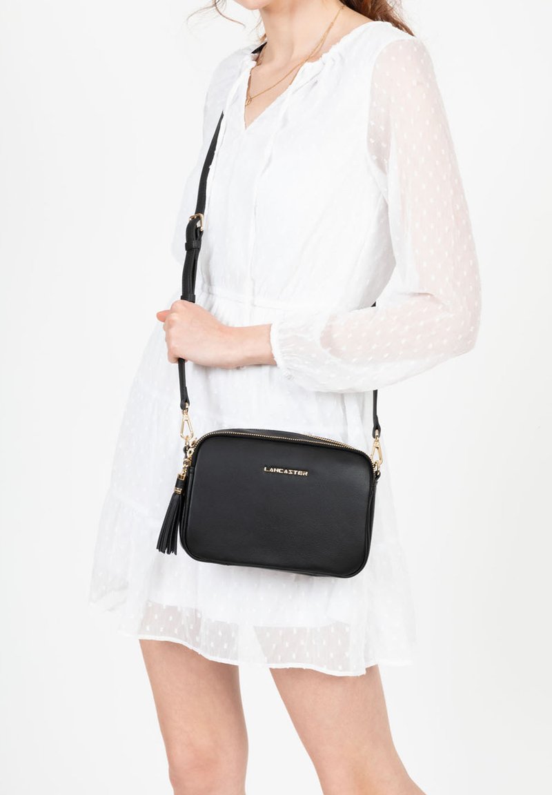 Borsa a tracolla in pelle nera con dettagli in oro, chiusura con zip e accentuazione a nappina. Texture liscia, forma rettangolare e logo del marchio visibile.