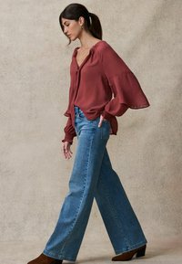 Langärmlige, rostfarbene Bluse mit rüschengeschnittenem Ausschnitt und weiten Ärmeln, kombiniert mit hellblauen geraden Jeans und braunen Stiefeletten mit Absatz.