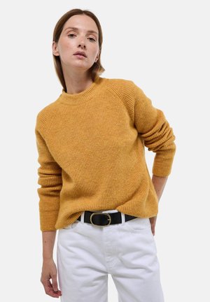 Pull en maille jaune moutarde avec une texture côtelée et des manches raglan, associé à un pantalon blanc et une ceinture noire avec une boucle en or.