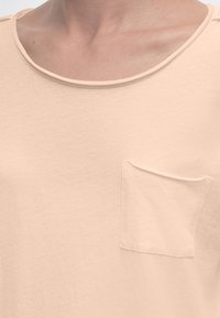 Φωτεινή μπεζ μπλούζα με στρογγυλή λαιμόκοψη και μικρό τσεπάκι στο αριστερό μέρος του στήθους, φορεμένη από πρόσωπο ορατό από το λαιμό μέχρι το στήθος.