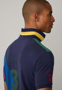Polo shirt van marineblauwe stof met meerkleurige accenten, kraag in geel en groen, geborduurd "HACKETT" en nummer 3 op de achterkant.
