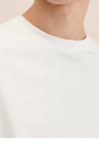 Bluza crewneck z białej bawełnianej tkaniny; posiada teksturowaną powierzchnię i luźny krój z rękawami raglanowymi. Brak widocznych wzorów ani logo.