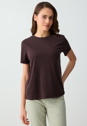 Jonge vrouw met een lichte huid en middenbruin haar draagt een donkerbruine korte mouw t-shirt en lichtbeige broek, staand tegen een effen achtergrond.