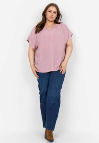 Blusa rosa chiaro a maniche corte con scollatura a V, abbinata a jeans blu a gamba dritta e stivali alla caviglia marroni con tacco.
