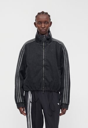 Person trägt eine schwarze Zip-up-Jacke mit drei grauen Streifen an den Ärmeln und schwarze Hosen mit weißen Seitenstreifen vor einem einfarbigen Hintergrund.