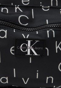 Μαύρη τσάντα υφάσματος με επαναλαμβανόμενο λευκό μοτίβο λογότυπου "Calvin Klein", με εξέχουσα κεντημένη ετικέτα που αναγράφει "CK Calvin Klein Jeans."