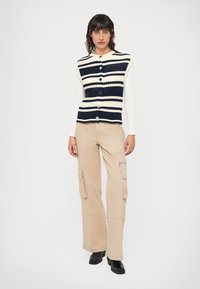 Colete de malha listrado em azul-marinho e creme com botões, combinado com calças cargo largas em bege claro com bolsos laterais. Botas de salto pretas.