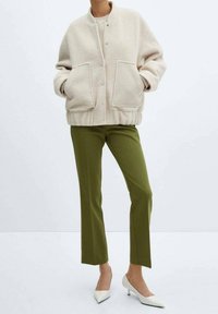 Persoon in een crèmewit textuur oversized jasje, olijfgroen op maat gemaakte broek en witte puntige lage hakken, staand op een witte achtergrond.