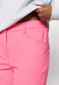 Rozā bikses, izgatavotas no gludas auduma, ar plakanu priekšpusi, sānu kabatām un taisna kāju dizainu. Izceltas ar minimālām šuvju detaļām.