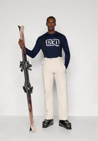 Beżowe spodnie narciarskie z regulowanymi szelkami, połączone z granatową koszulą z długim rękawem, na której znajduje się biały napis „SKI”, trzymające narty z wiązaniami.