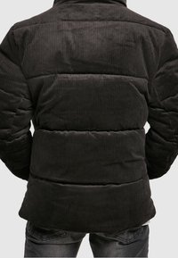 Černá manšestrová péřová bunda s prošívaným vzorem, žebrovanou texturou a vysokým límcem. Zadní pohled ukazuje stažený pás a detaily vycpávek.